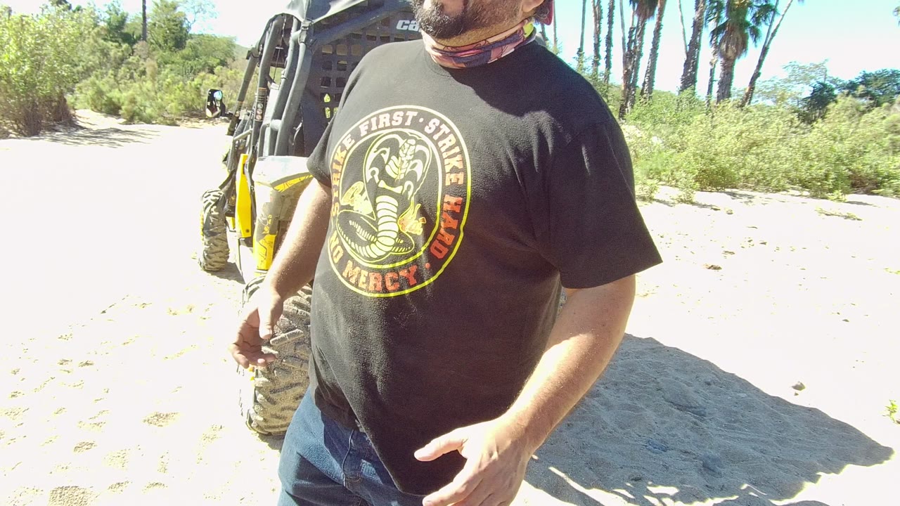 In Los Cabos, the man stands beside the yellow-accented off-road vehicle and begins gesturing with his hand over the sunlit sand and scrubby background.