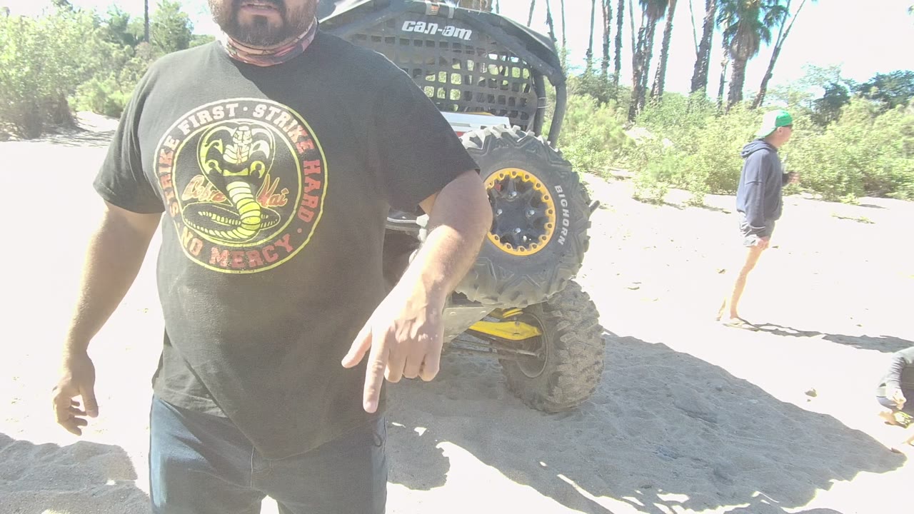 In Los Cabos, a cropped man in a black Cobra Kai T-shirt stands close to camera on sandy ground while an off-road vehicle tire and background walker are visible.