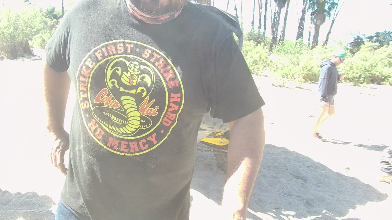 In Los Cabos, the foreground man's Cobra Kai shirt fills more of the frame as a Can-Am off-road vehicle and a person in the background remain visible in bright light.