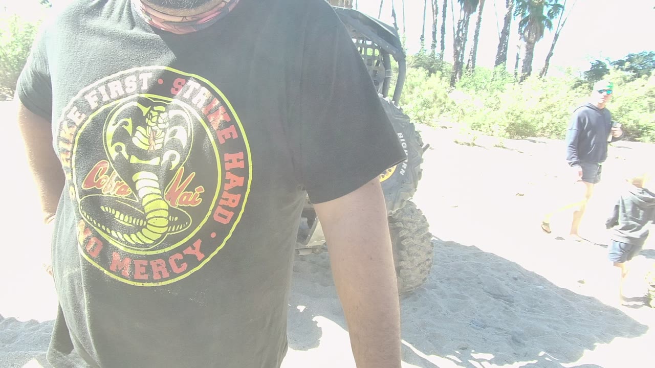 In Los Cabos, the cropped man remains close to camera with the Cobra Kai shirt centered, while people walk across the bright sandy background near the off-road vehicle.