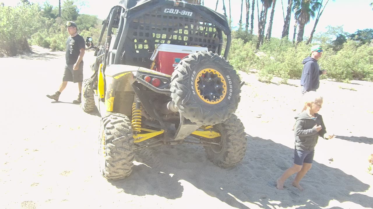 Rear view of a yellow Can-Am off-road vehicle on sandy ground in Los Cabos, with an adult walking left and a child near the vehicle.