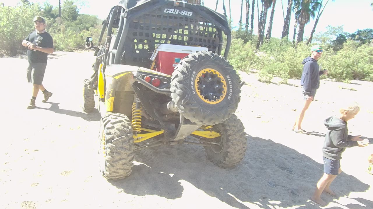 People move around a parked yellow off-road vehicle on bright sand.