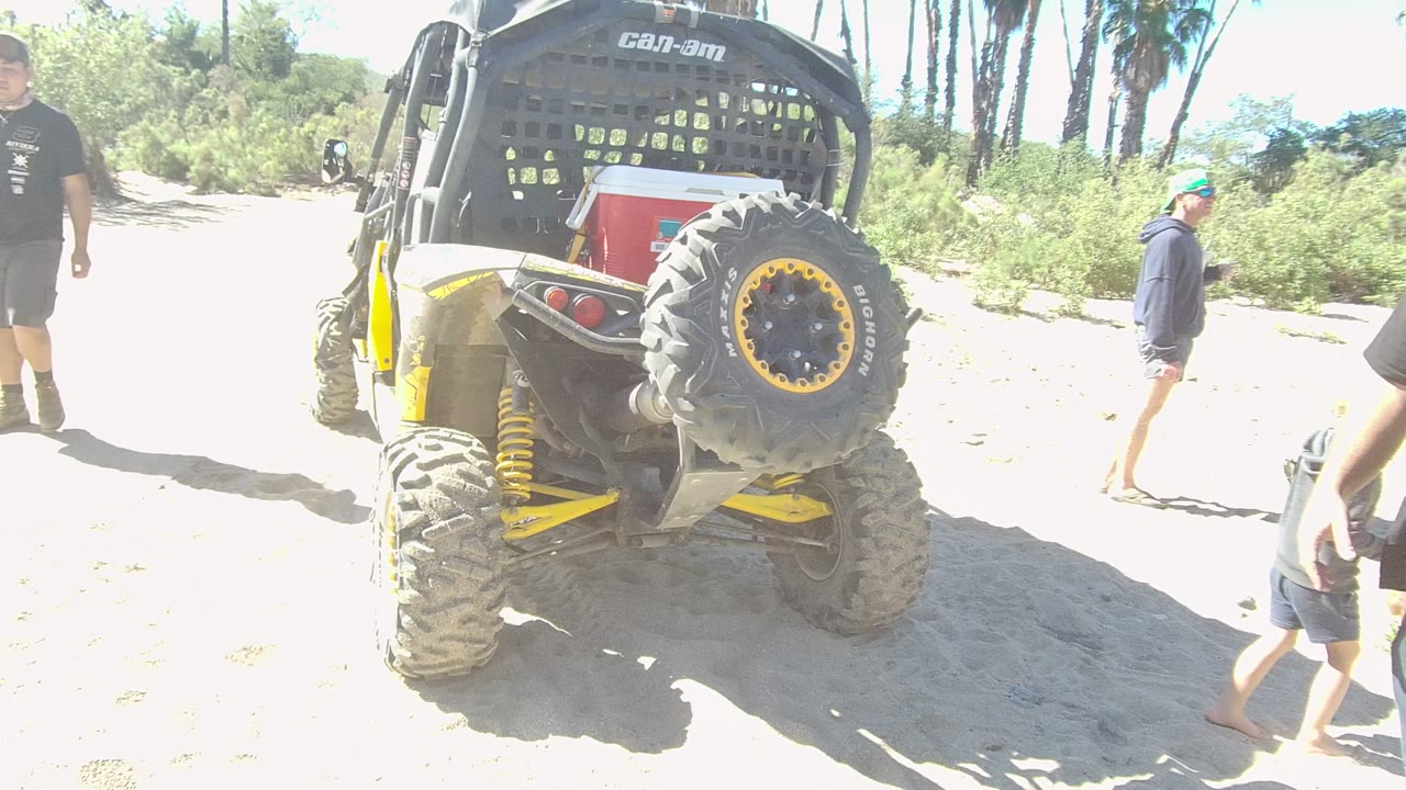 At two seconds, people continue crossing near the rear of the yellow Can-Am off-road vehicle on sandy Los Cabos terrain, with palms in the background.