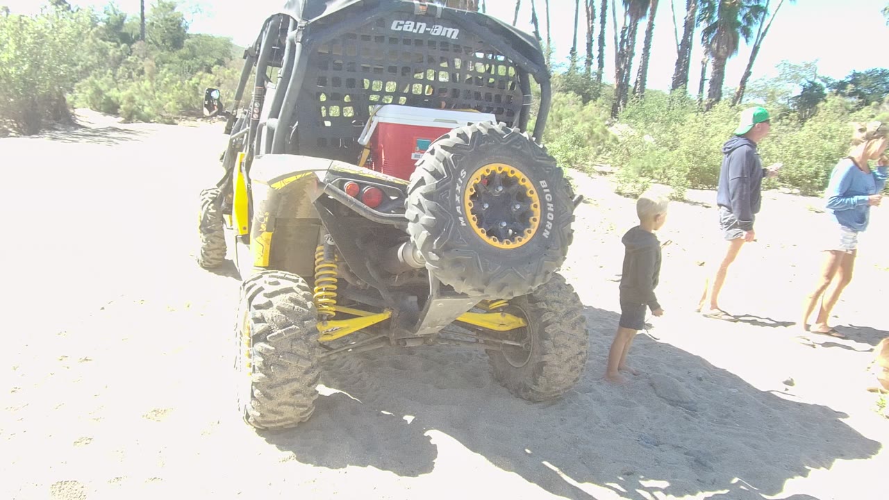 In Los Cabos, Mexico, a yellow and black dune buggy sits on bright sand as a child stands near the rear tire and two adults walk in the background.