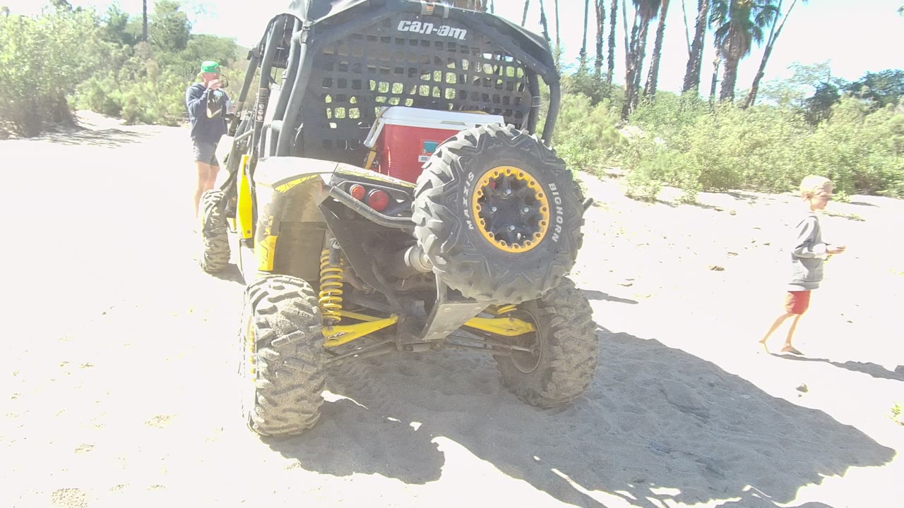 In Los Cabos, Baja California Sur, Mexico, a yellow and black Can-Am UTV is parked on sand while an adult and a child are visible near the vehicle.