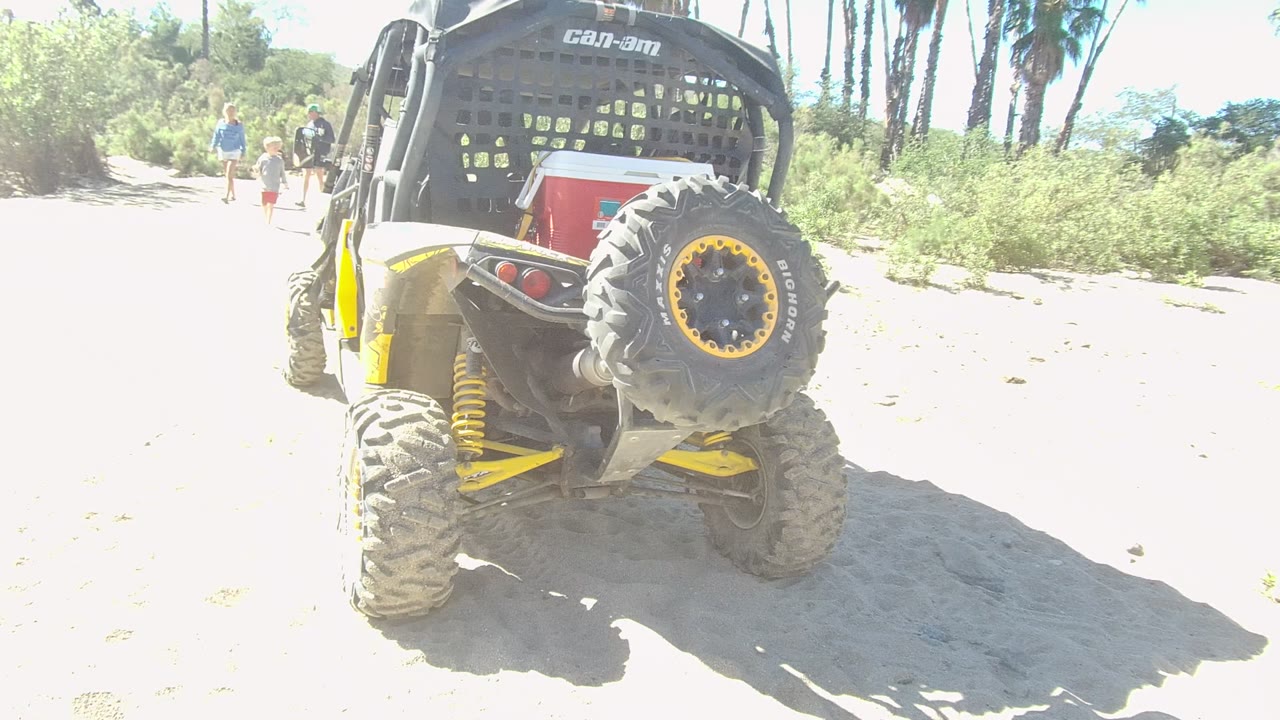At 0:00 in Los Cabos, Baja California Sur, a yellow and black Can-Am UTV sits close to camera on sandy ground while distant people walk along a bright path.