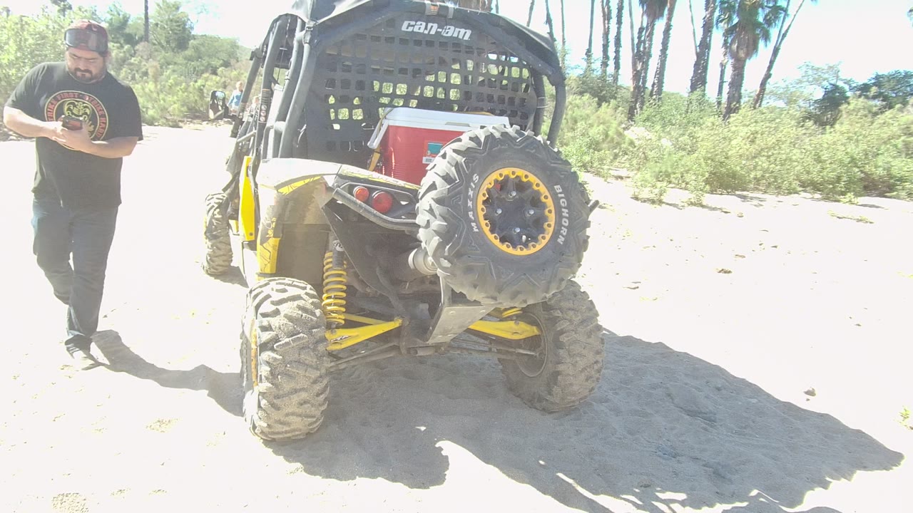 In Los Cabos, Mexico, a man in a black T-shirt and cap walks near the rear of a yellow and black off-road vehicle on a bright sandy path.