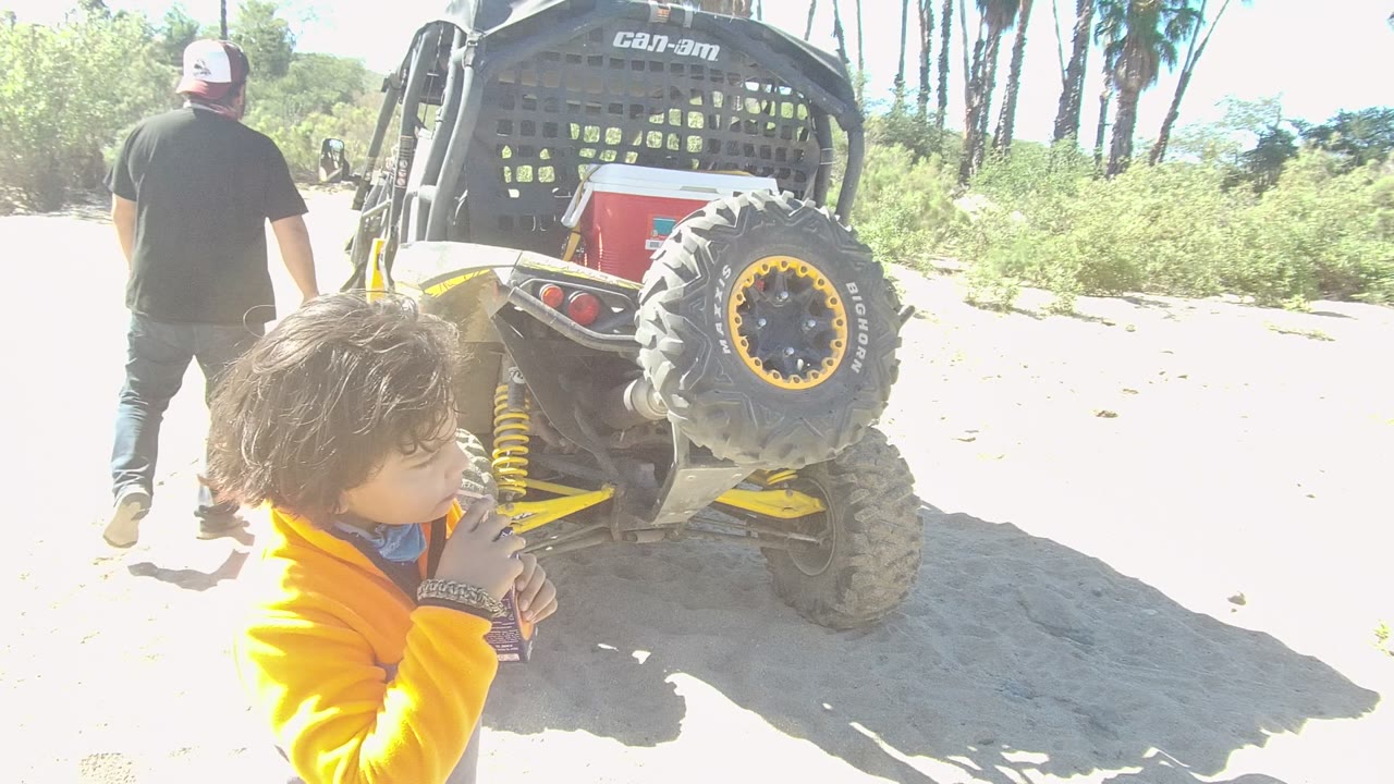In Los Cabos, Baja California Sur, a child in a yellow jacket stands close to the rear of a yellow and black off-road vehicle while a man walks away on sandy terrain.