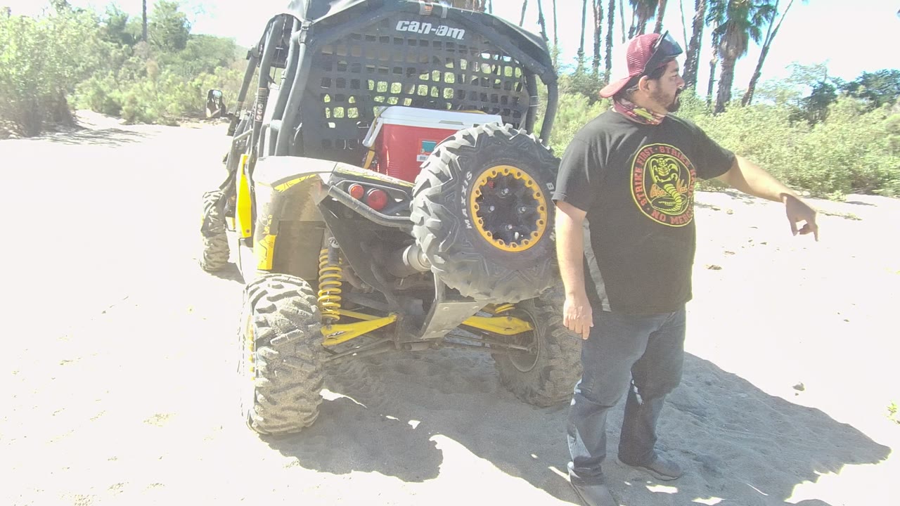 In Los Cabos, Baja California Sur, a man in a black shirt and red cap points beside the rear of an off-road UTV with a spare tire on bright sandy ground.