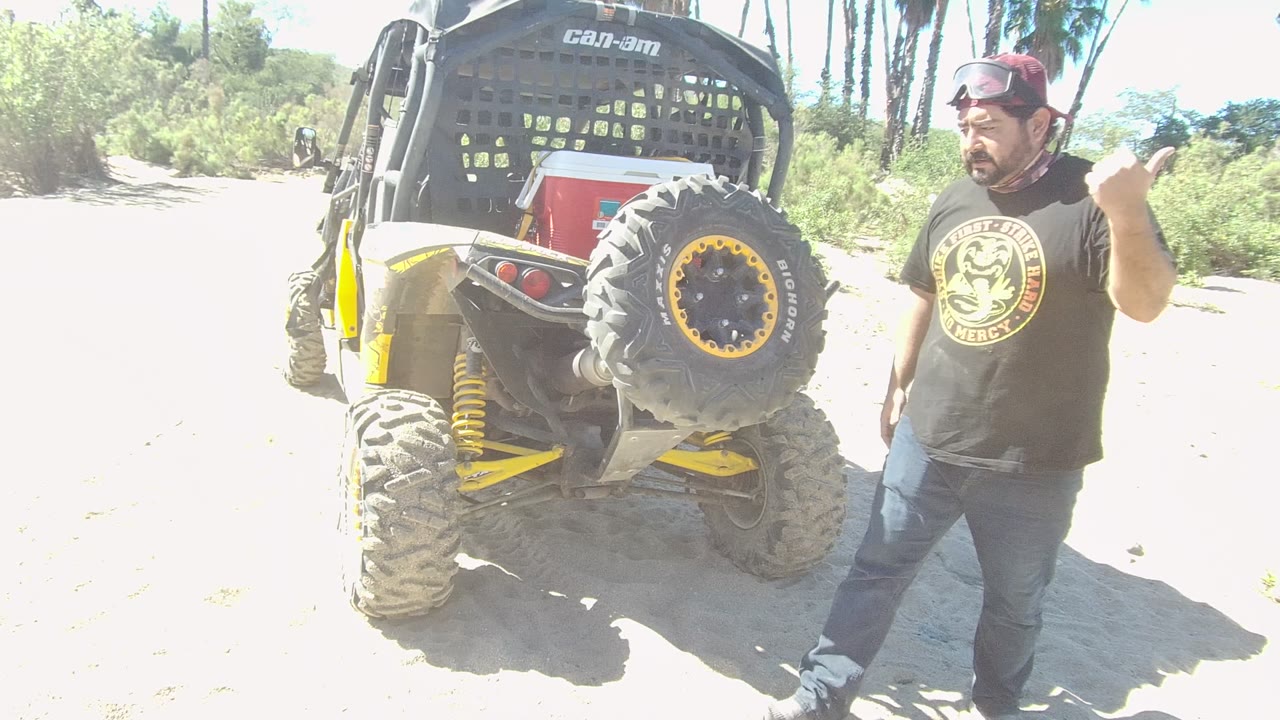 The man raises one hand while standing beside the rear of the UTV in Los Cabos, with the vehicle's spare tire and roll cage prominent on the left.