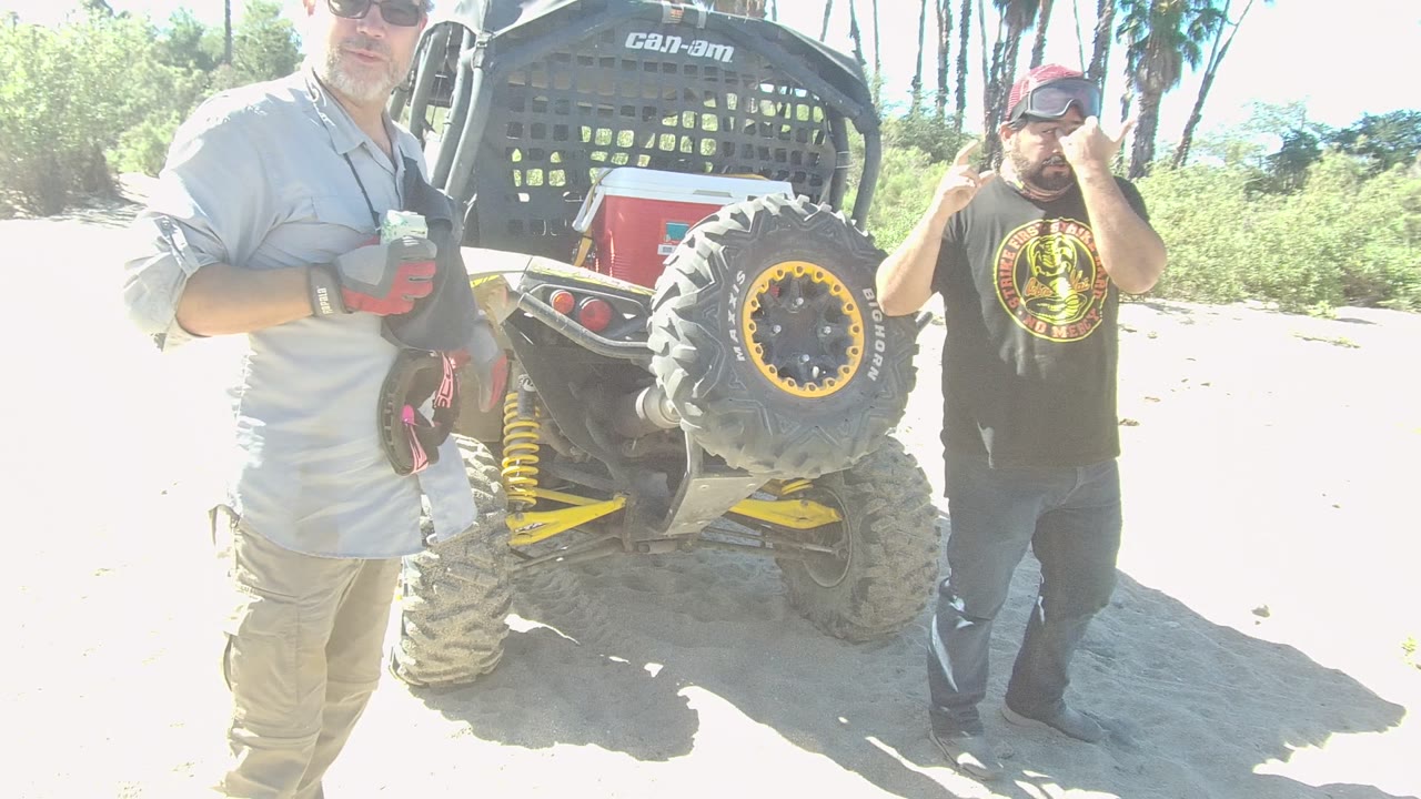 Two adult men stand beside the rear of a Can-Am off-road vehicle on sandy ground in Los Cabos, with one man in sunglasses near the camera and another adjusting his hat.