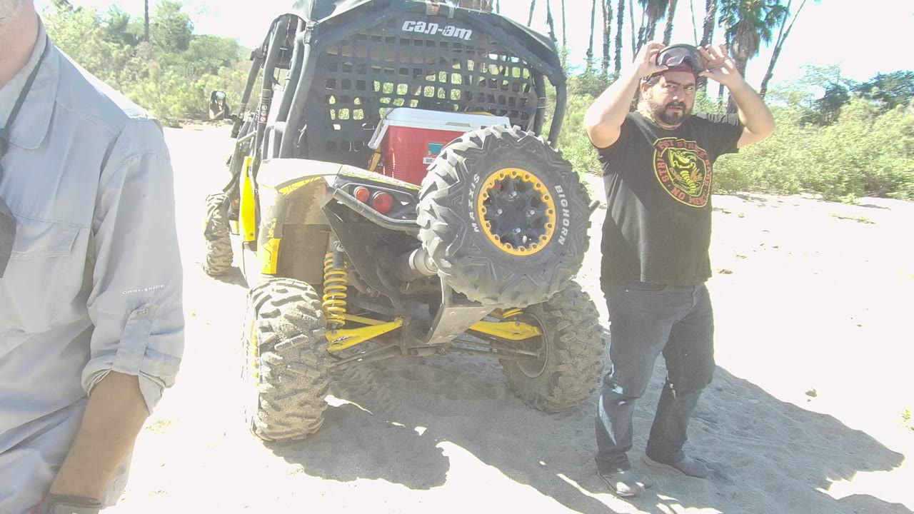 The man in a black T-shirt gestures with one hand beside the rear of the Can-Am off-road vehicle in Los Cabos as the other man moves out of the frame.