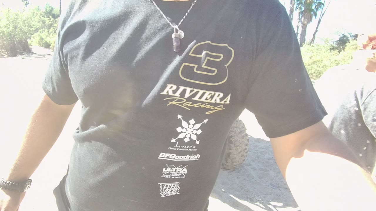 Close foreground view of a person wearing a black Riviera Racing T-shirt in Los Cabos, with sandy ground and bright outdoor light behind the cropped body.