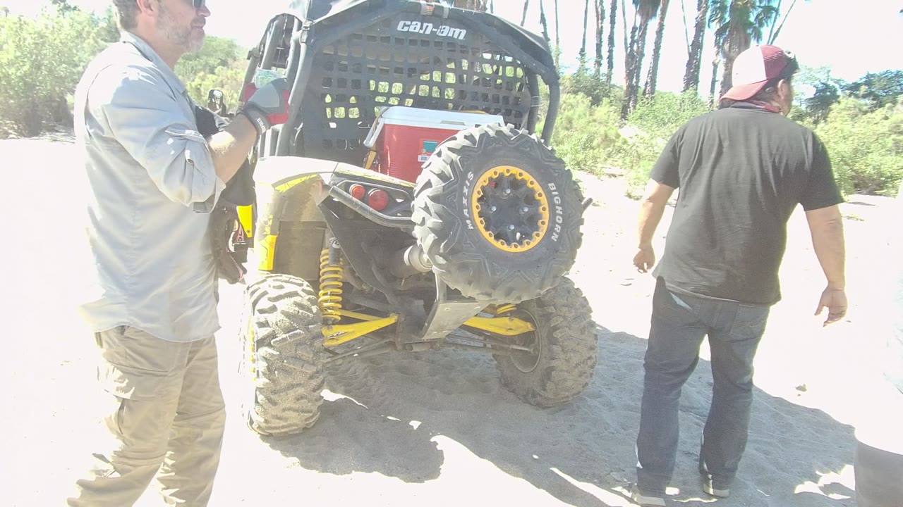 A man in a light shirt holds a camera beside the rear of a Can-Am off-road vehicle in Los Cabos while another man in a black shirt walks nearby on sand.