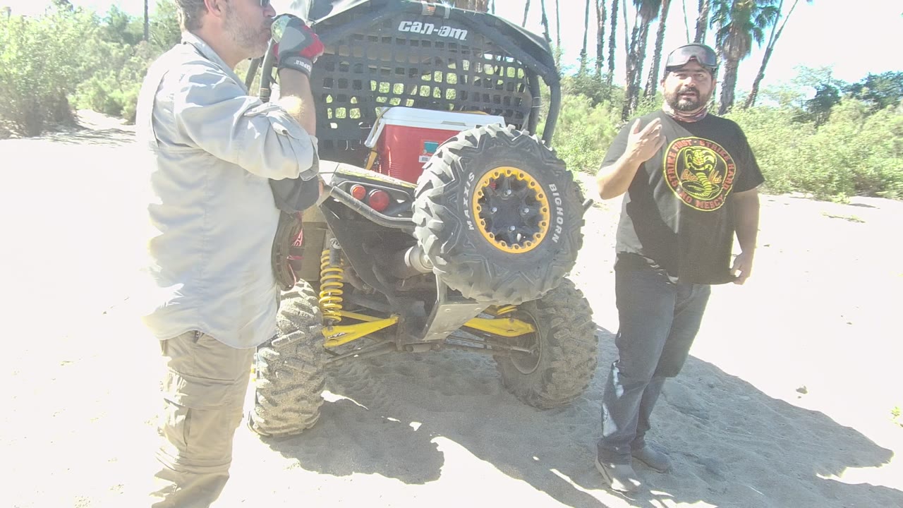 At 0:00 in Los Cabos, Baja California Sur, two adult men stand near the rear tire and cargo rack of a black Can-Am dune buggy on bright sandy terrain.