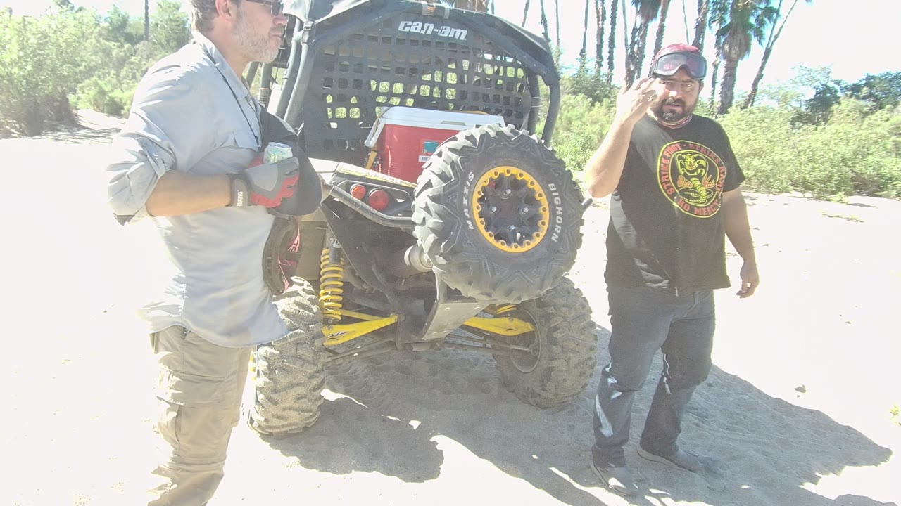 Man in black shirt gestures near a dune buggy while another man stands cropped in foreground.