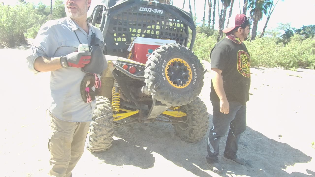 At 0:02 in Los Cabos, Baja California Sur, the man in the light shirt faces left holding gear while the man in the black shirt turns slightly beside the Can-Am buggy.