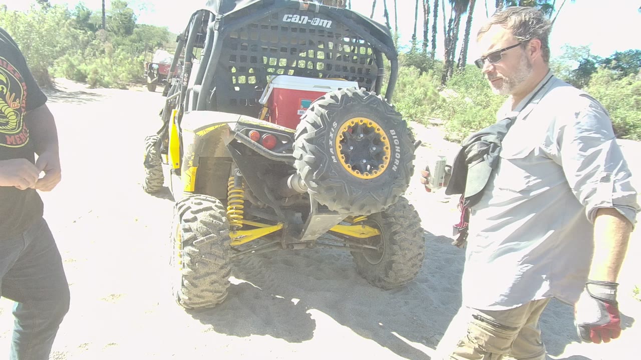 In Los Cabos, two adult men pass near the camera while a yellow and black Can-Am utility vehicle is parked on a bright sandy trail with palms behind it.