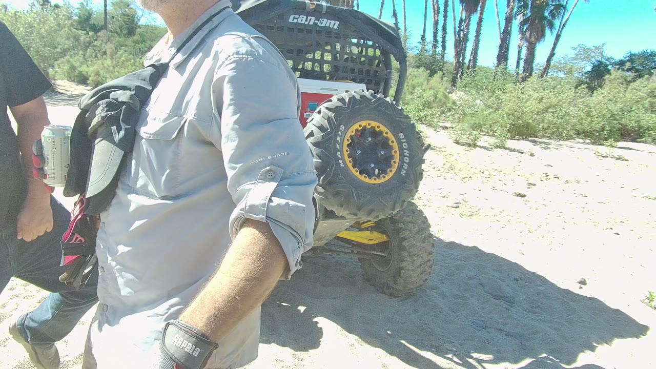 In Los Cabos, the frame crops close to the torso of a man in a light shirt while another person passes at left and the parked Can-Am UTV fills the right side of the sandy trail scene.