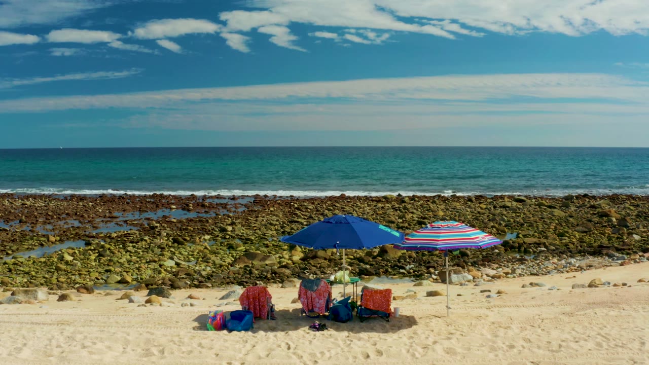 At El Tule Los Cabos, two umbrellas and red beach chairs sit on pale sand below a rocky shoreline and turquoise ocean under a partly cloudy sky, with no visible people.