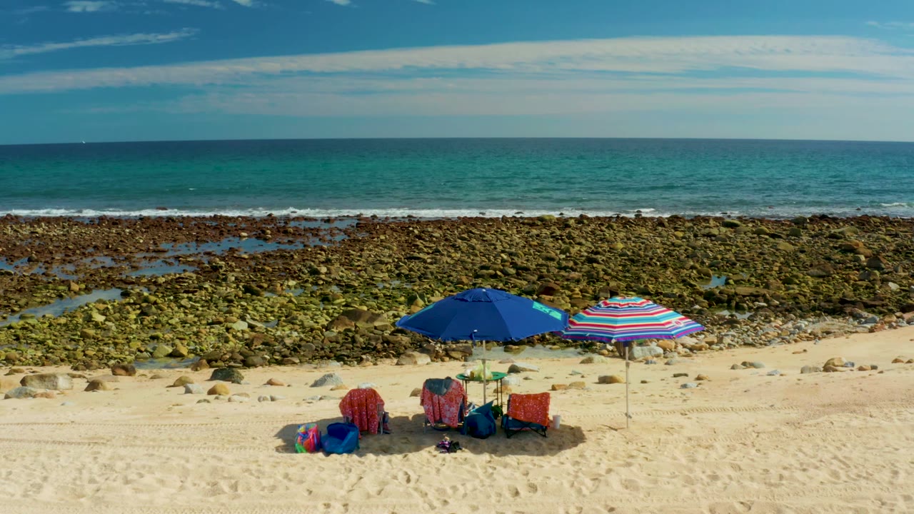 Umbrellas and chairs on sand beside rocky tide pools and blue ocean.