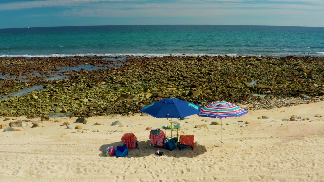 At El Tule Los Cabos, two umbrellas, red chairs, and bags sit on the sand at the lower right of a wide view of exposed rocks, shallow pools, and ocean, with no visible people.