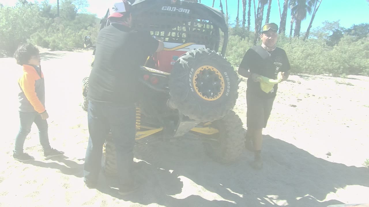 In Los Cabos, a man in black stands by the rear of a red off-road vehicle while a child in orange and another adult are visible on the sandy track.