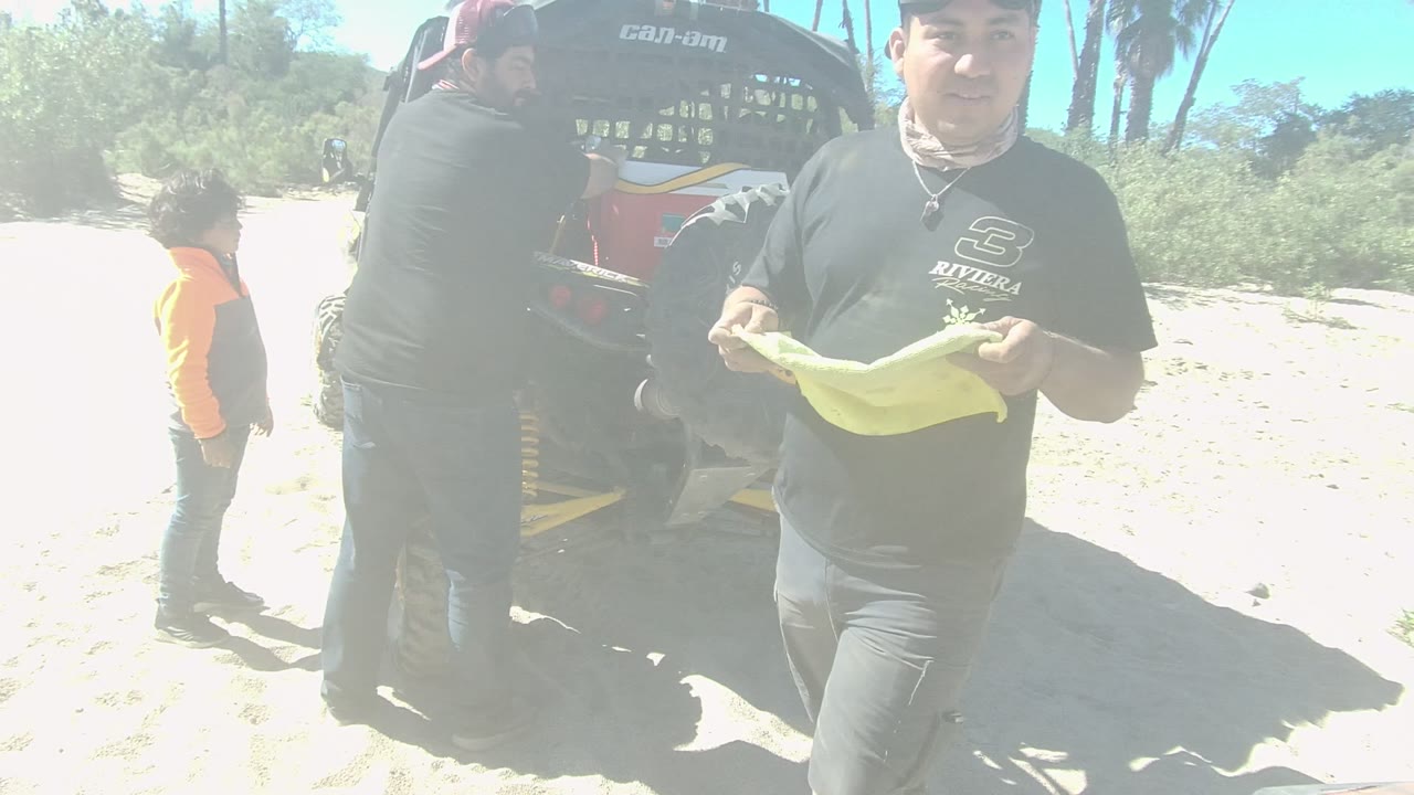 The man with the yellow towel steps nearer to the camera in Los Cabos, with the off-road vehicle partly behind him and a child standing to the side.