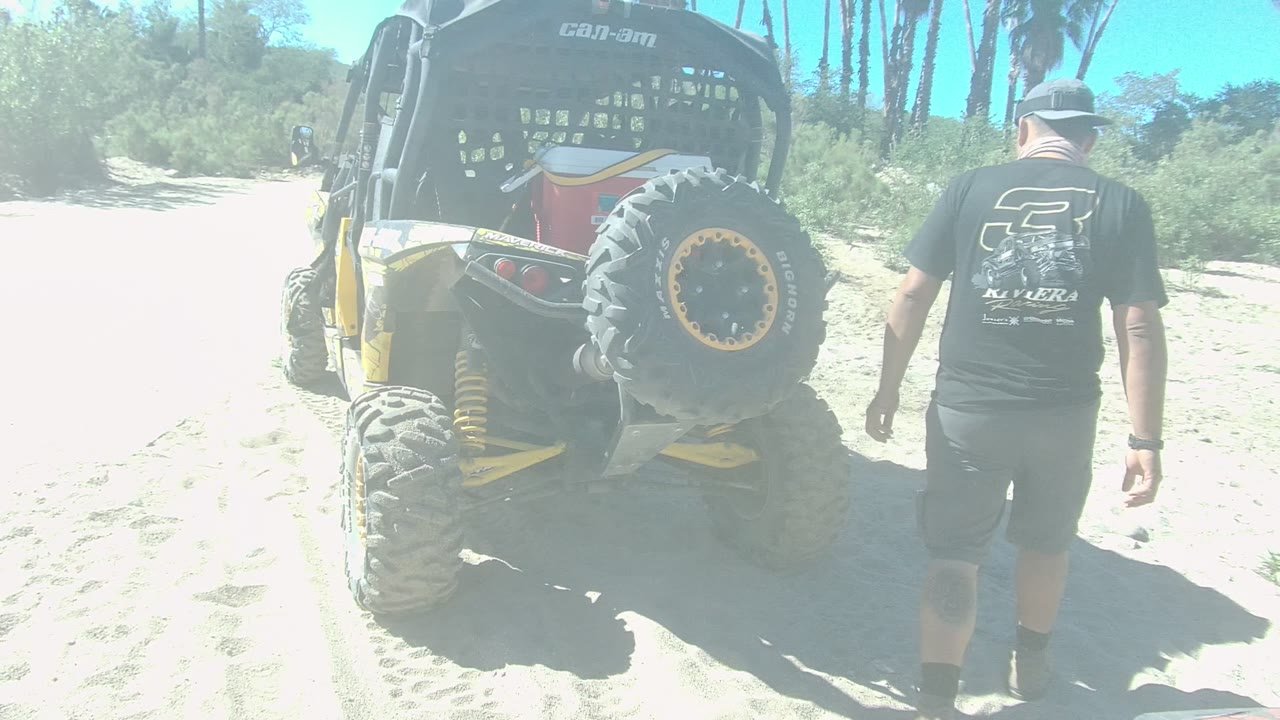 In Los Cabos, Baja California Sur, Mexico, a man in a black shirt and cap walks toward the rear of a parked yellow and black dune buggy on bright sand.