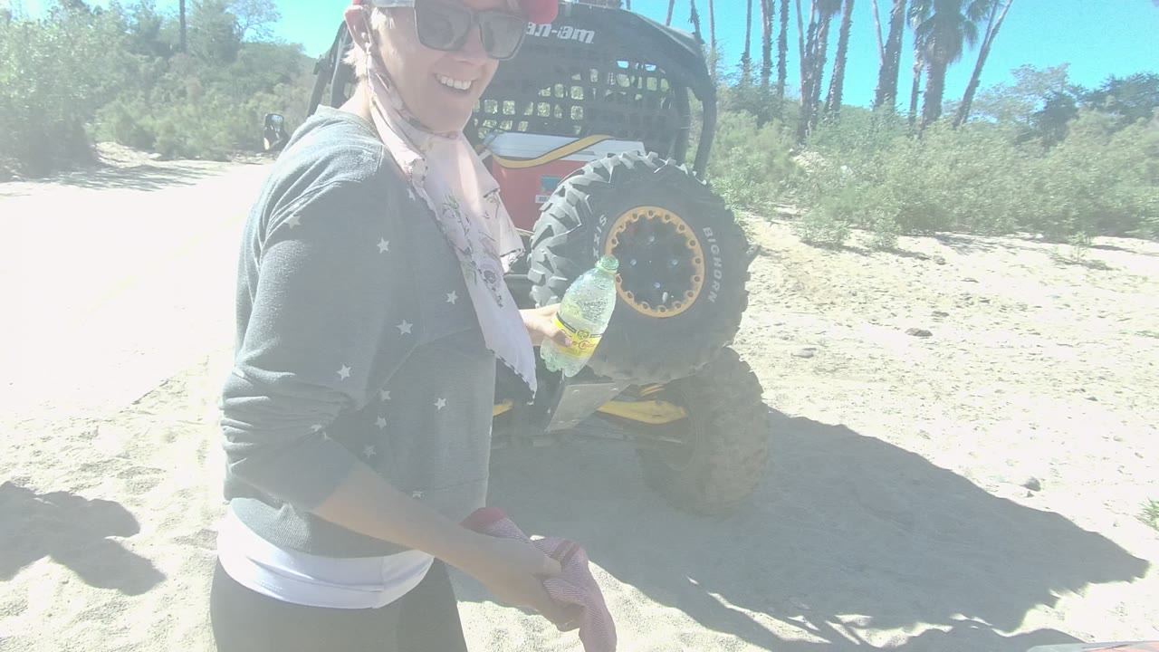 In Los Cabos, Baja California Sur, a person in sunglasses and a red cap stands beside a dune buggy while holding a bottle on bright sand.
