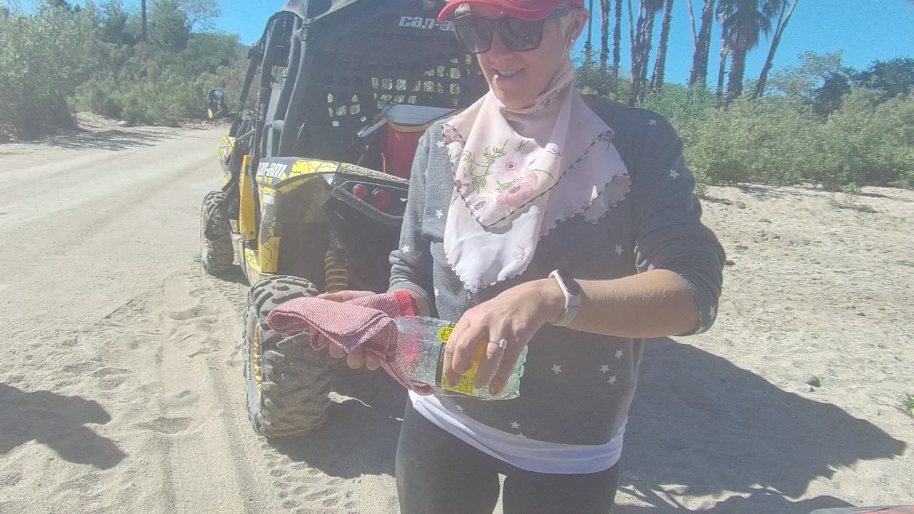 In Los Cabos, Baja California Sur, the person stands closer to the lens with a bottle and cloth, with the dune buggy rear and sandy track behind them.