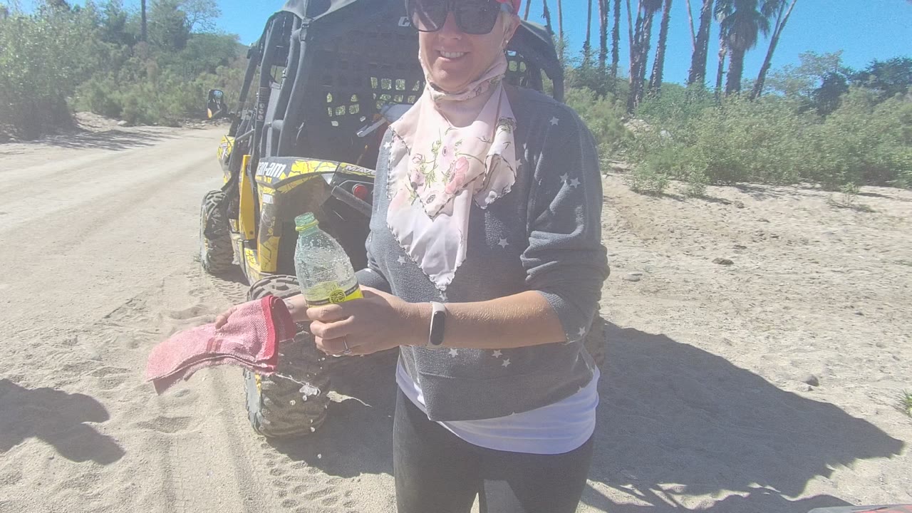 In Los Cabos, Baja California Sur, the person smiles toward the camera while holding a bottle and pink cloth beside the parked dune buggy.