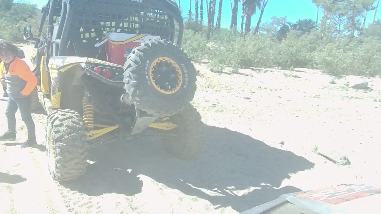 Rear of an off-road vehicle in Los Cabos with a child in an orange shirt at the left edge and another person near sandy tracks under bright sun.