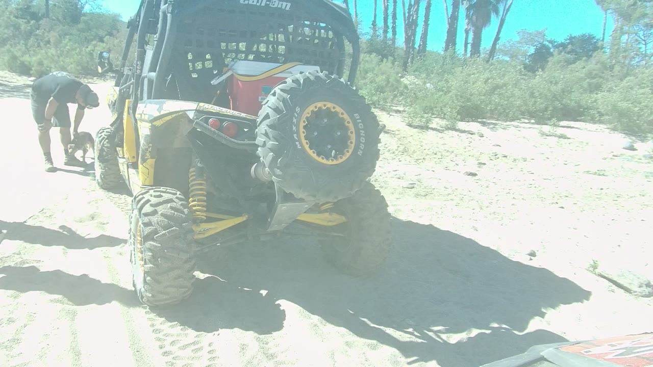 The rear tire and suspension of a Can-Am off-road vehicle dominate the left side while people work nearby on sandy ground in Los Cabos.