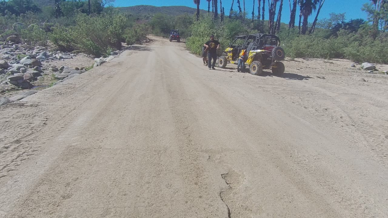 Wide view of a sandy road in Los Cabos, Baja California Sur, with a yellow UTV parked or moving near people on the right and scrub-covered desert terrain in the background.