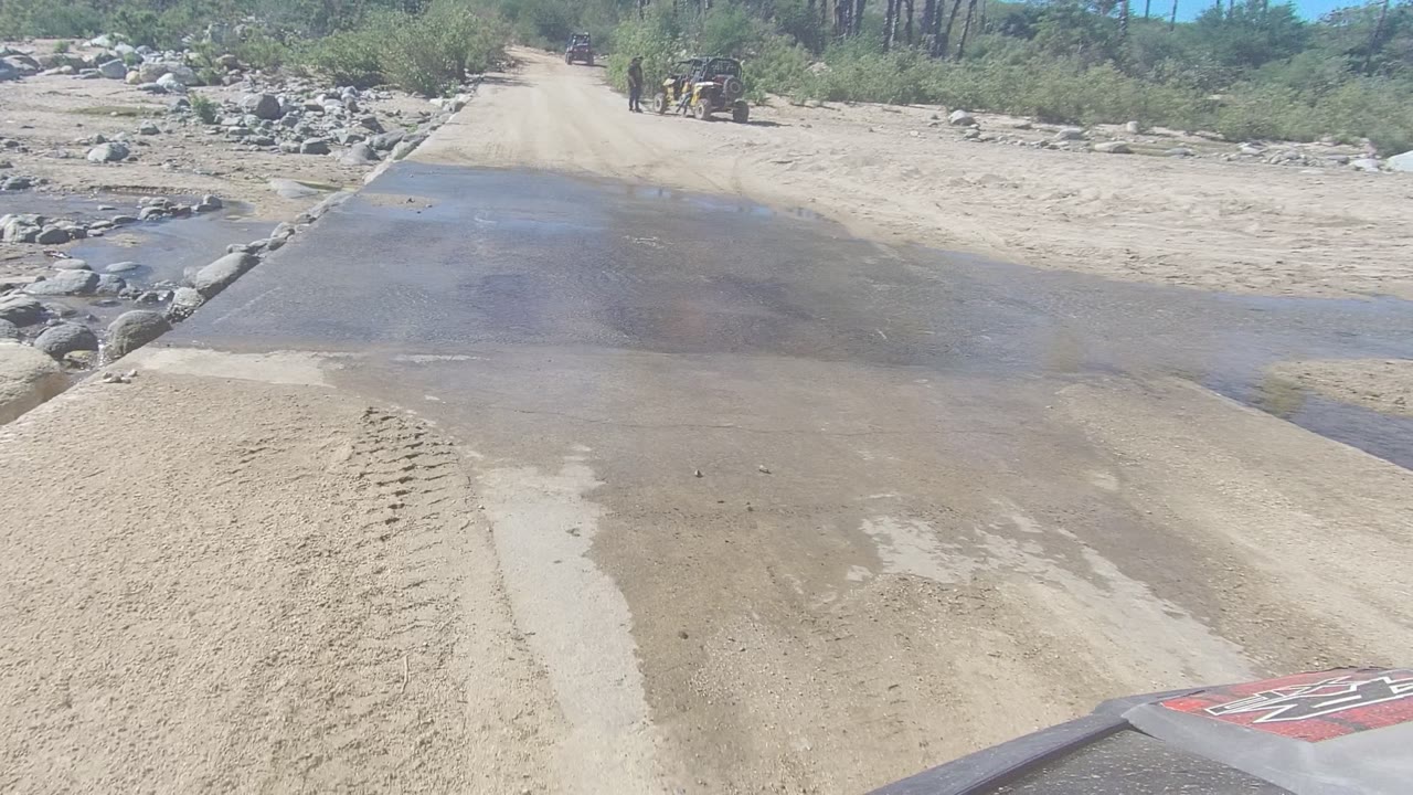 At 0:00 in Los Cabos, Baja California Sur, shallow water covers the foreground of a sandy dirt road, with rocks, scrub vegetation, and a distant off-road vehicle near the upper road.