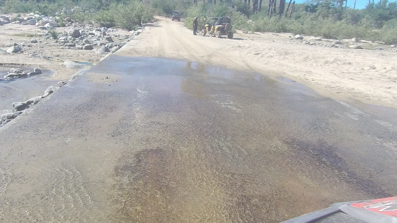 Dirt road water crossing with receding water and off-road vehicles in the background.