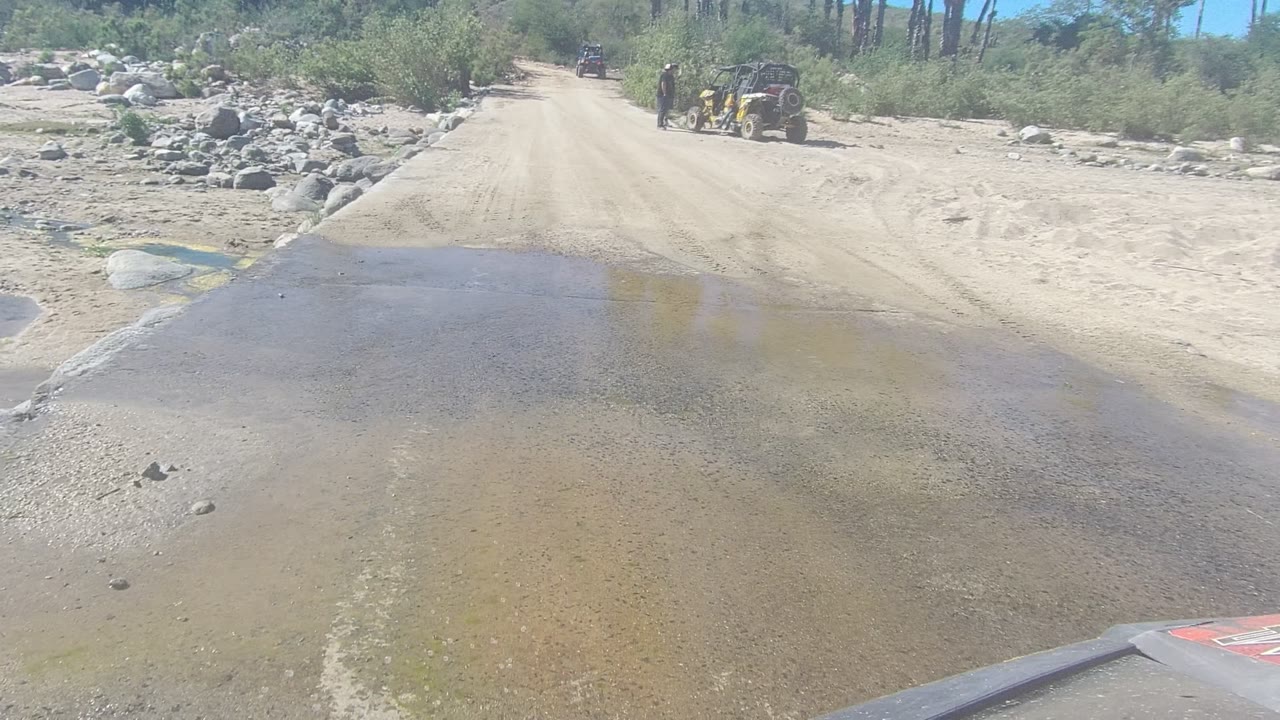 At 0:02 in Los Cabos, Baja California Sur, shallow water thins across the road while a yellow UTV and a standing person are visible near the right side of the distant track.