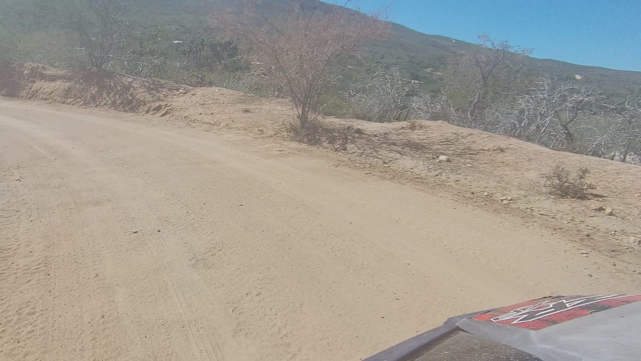 Start of the Los Cabos clip shows a sandy unpaved road beside dry shrubs and low hills, with part of the vehicle hood visible and no visible people.