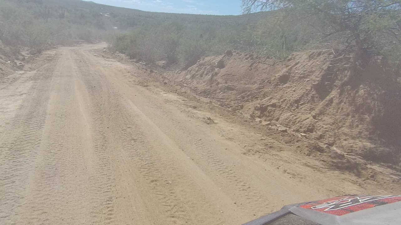 At 0:02, the Los Cabos dirt road stays centered as dusty air softens the view of scrub, roadside cuts, and low hills, with no visible people.