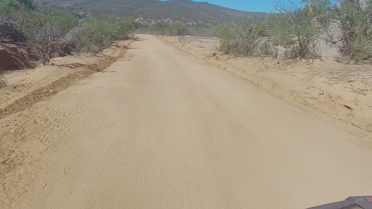 Opening view of a sandy dirt road in Los Cabos, Baja California Sur, with scrubby bushes on both sides, low hills in the distance, and no visible people.