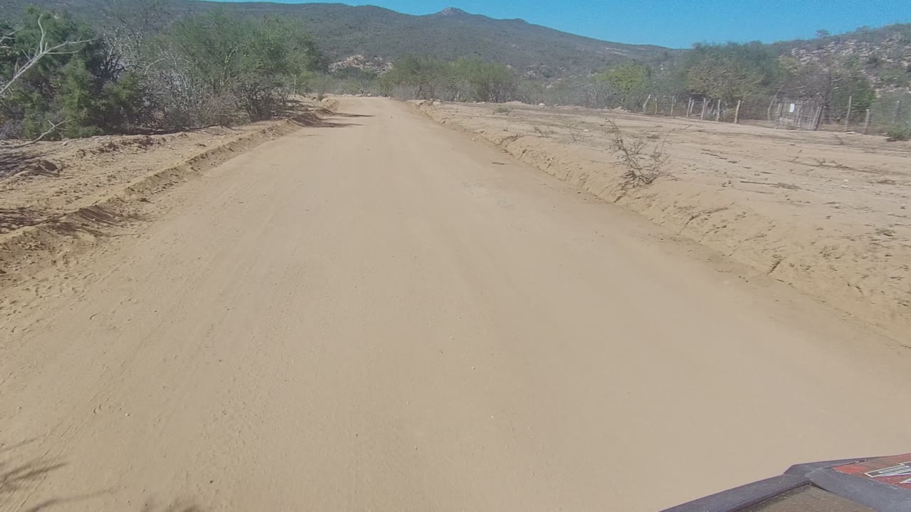 The road view continues in Los Cabos, Baja California Sur, with a broad tan surface, scrub vegetation to the left and right, distant hills, and no visible people.