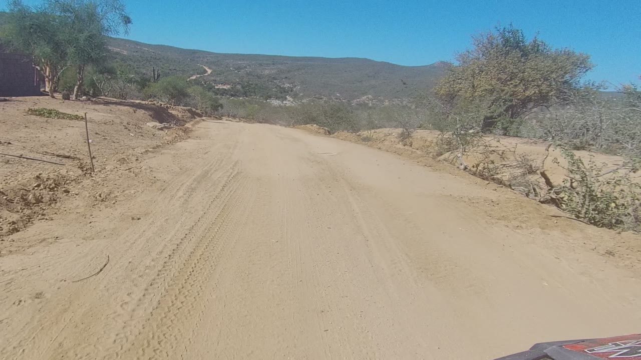 A forward-facing view in Los Cabos shows a sandy road bordered by scrub and a raised eroded bank, with low hills beyond and no visible people.