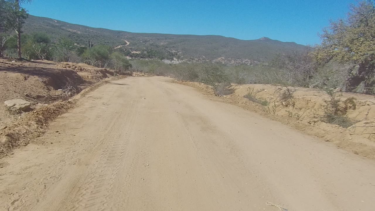 Wide dirt road descending through desert-like brush toward hills.