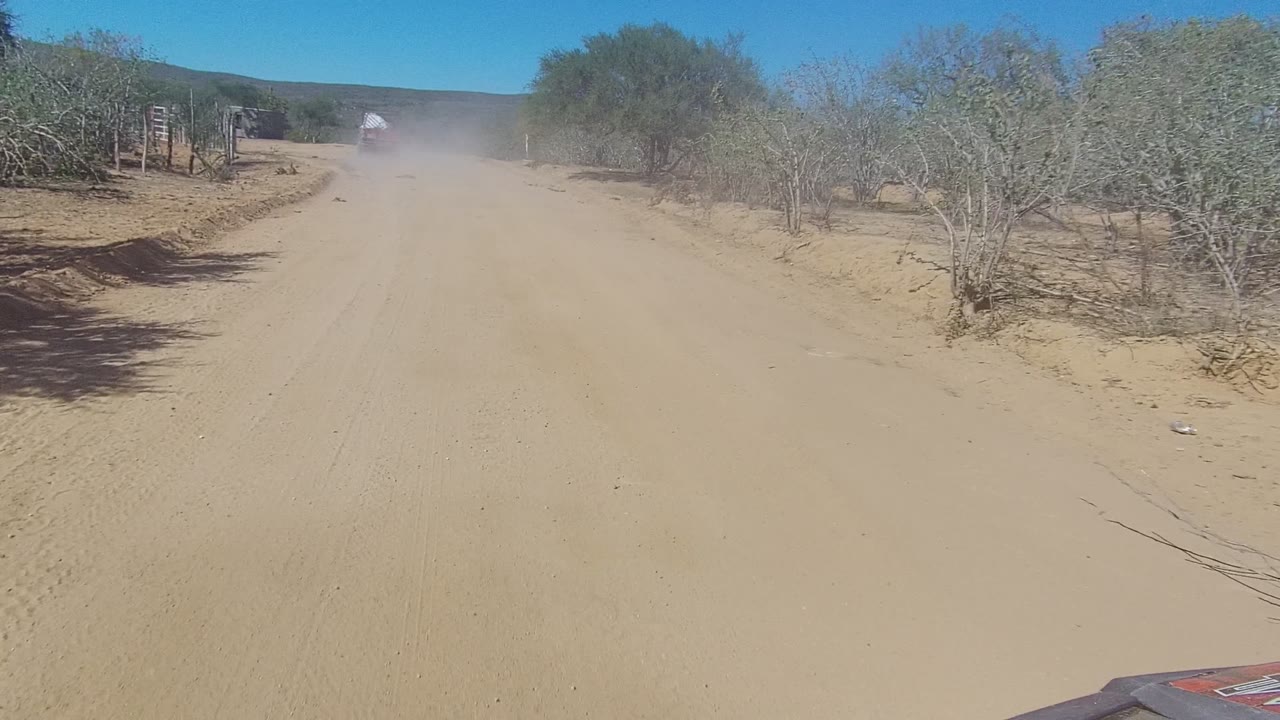 Dusty dirt road passing dry brush and a simple roadside structure.