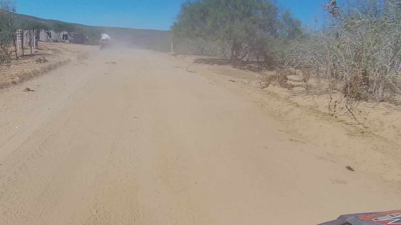 Wide Los Cabos sandy road frame with scrub vegetation on both sides, a dust plume farther ahead, clear blue sky, and no visible people.