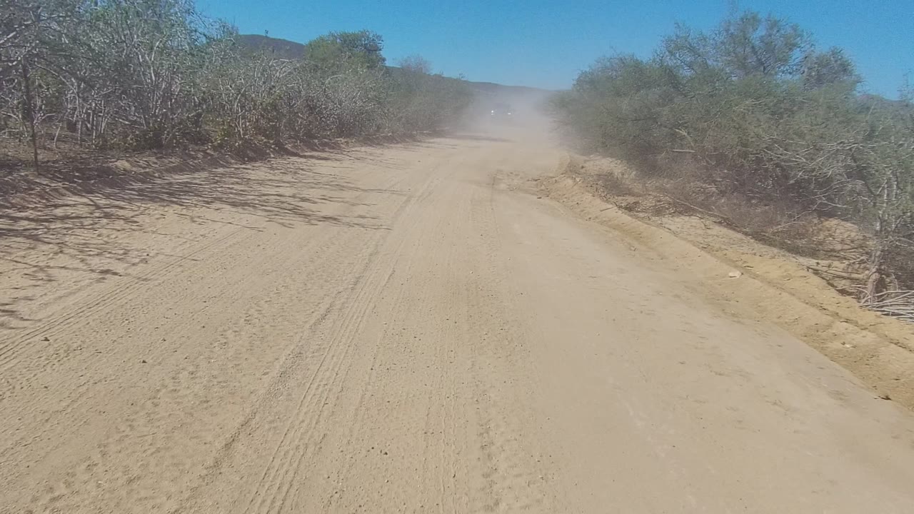 A sandy dirt road in Los Cabos, Baja California Sur runs between scrub vegetation as a distant truck approaches through dust, with no visible people.