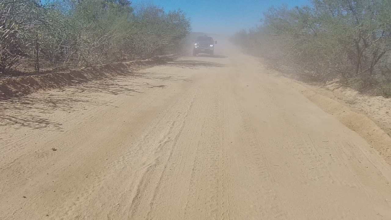 A dark pickup truck approaches on the Los Cabos dirt road, framed by dry brush and a lingering dust plume under a clear blue sky, with no visible people.