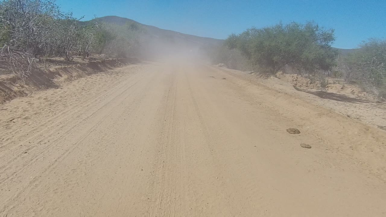 At 0:00, a sandy dirt road in Los Cabos, Baja California Sur runs forward between dry shrubs, with dust hanging near the distant road and no visible people.
