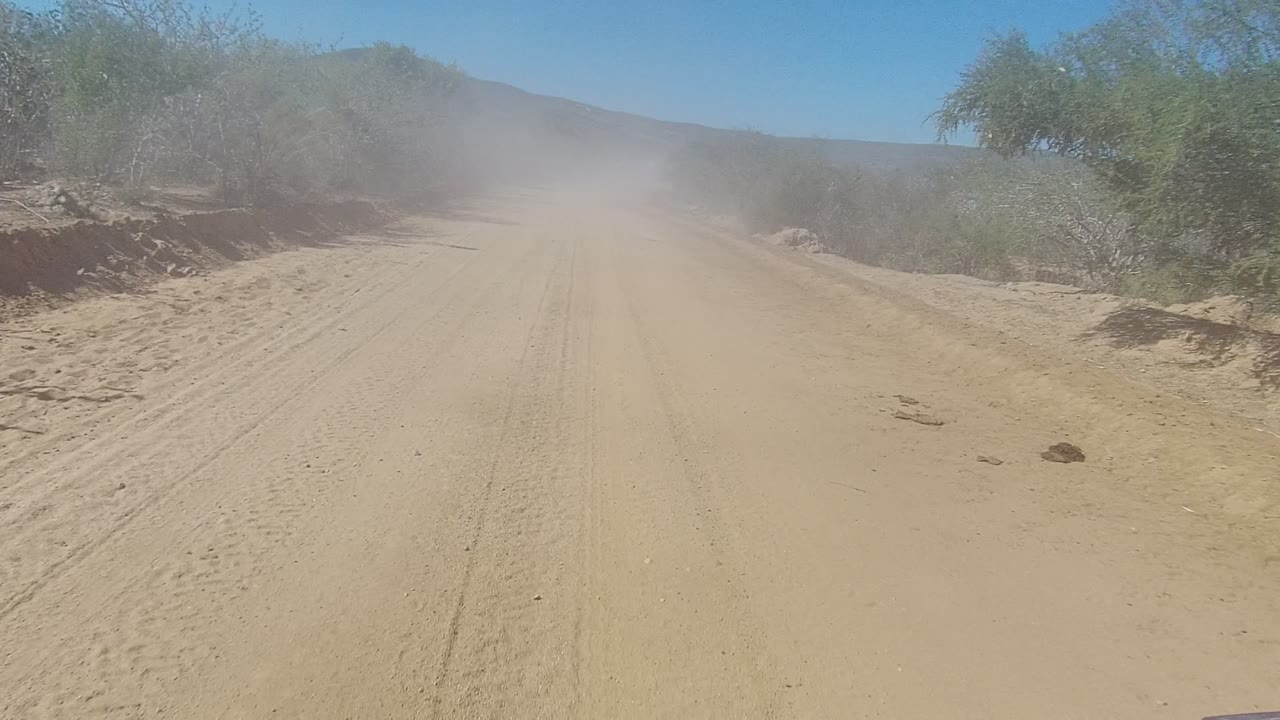 Wide dirt road with a dust plume drifting over shrubs and distant hills.